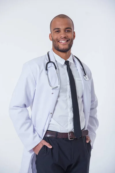 帅美国黑人穿白大褂的医生是看着相机 微笑而站在一起手在口袋里 在白色背景上孤立 — 图库照片