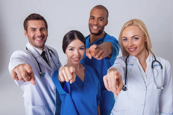 Group of medical doctors of different nationalities and genders is smiling, looking and pointing at camera, standing on gray background