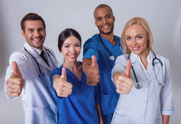 Group of medical doctors of different nationalities and genders is showing Ok sign, looking at camera and smiling, standing on gray background