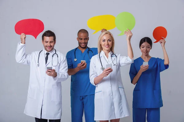 Group of medical doctors of different nationalities and genders is holding speech bubbles, using smartphones and smiling, standing on gray background