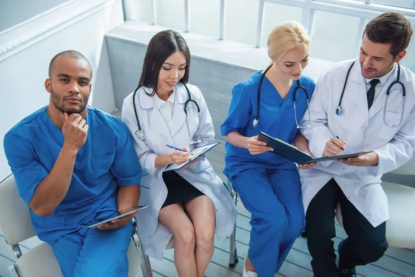 Group of medical doctors of different nationalities and genders with folders and tablet is examining documents and smiling, sitting in office. One is looking at camera