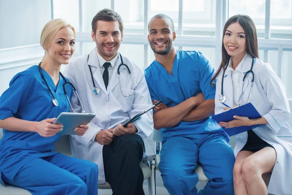 Group of medical doctors of different nationalities and genders with folders and tablet is looking at camera and smiling, sitting in office