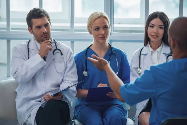 Medical doctors of different nationalities and genders are making notes while listening to another doctor\'s report, sitting in office