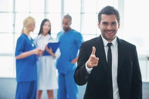 stock image Handsome man in classic suit is smiling, looking and pointing at camera, in the background three doctors are talking and making notes