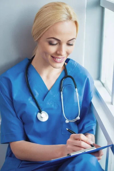 Piękna Blondynka Lekarka Niebieski Nosić Medycznych Jest Dokonywanie Notatek Uśmiechnięty — Zdjęcie stockowe