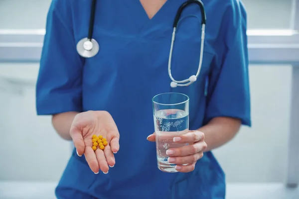 美丽的女医生的形象出现在蓝色医用服 拿着药和一杯水 — 图库照片