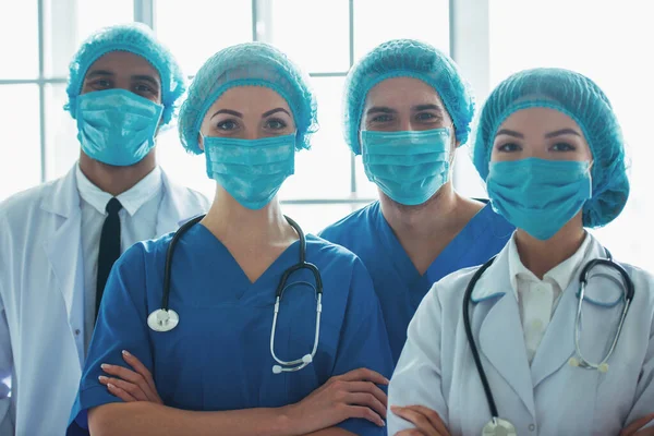Medical doctors of different nationalities and genders in masks and caps are looking at camera and smiling, standing with crossed arms