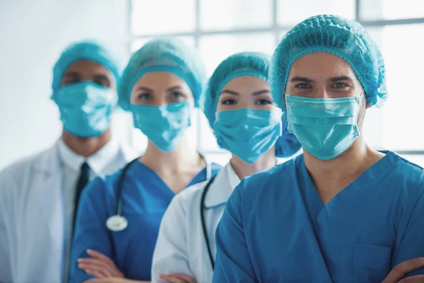 Medical doctors of different nationalities and genders in masks and caps are looking at camera, standing in a row one by one