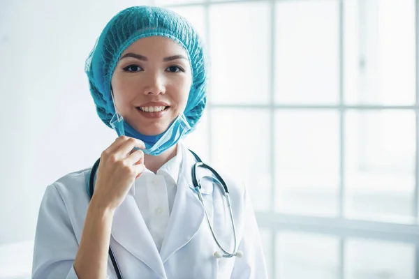 Beautiful Asian female doctor in cap is pulling mask, looking at camera and smiling