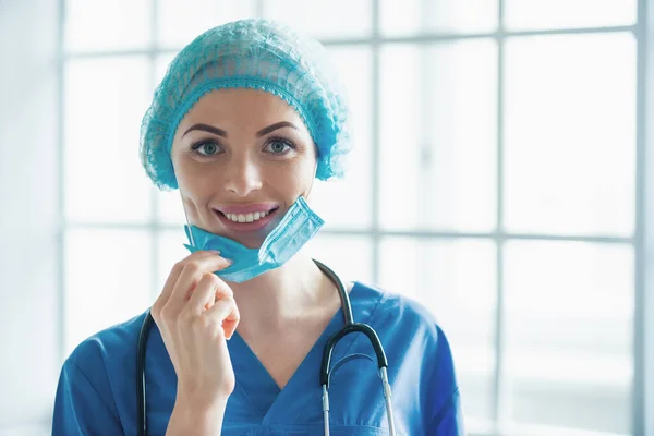 Beautiful female doctor in cap is pulling mask, looking at camera and smiling