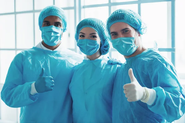 Surgeons of different nationalities and genders in masks and caps are looking at camera. Men are showing Ok sign