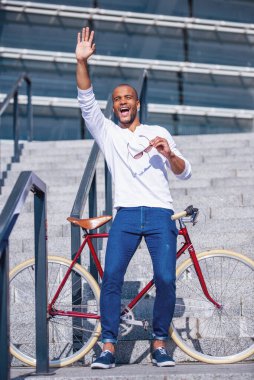Yakışıklı genç iş adamının gündelik kıyafetleriyle uzun bir portresi. El sallıyor, gülümsüyor, bisiklete yaslanıyor ve dışarıda merdivenlerde dikiliyor.