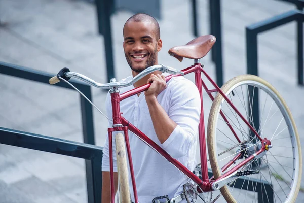 カジュアルな服を着たハンサムな若いビジネスマンは 自転車を持ちながらカメラを見て笑顔で 屋外に立っています — ストック写真