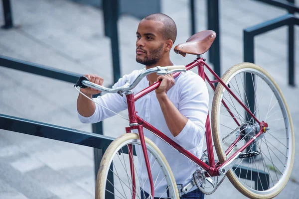 カジュアルな服を着たハンサムな若いビジネスマンは 自転車を持っている間 屋外に立って離れて見て考えています — ストック写真