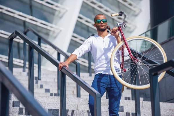 カジュアルな服やサングラスでハンサムな若いビジネスマンは 屋外の階段に立っている間に自転車を保持しています — ストック写真