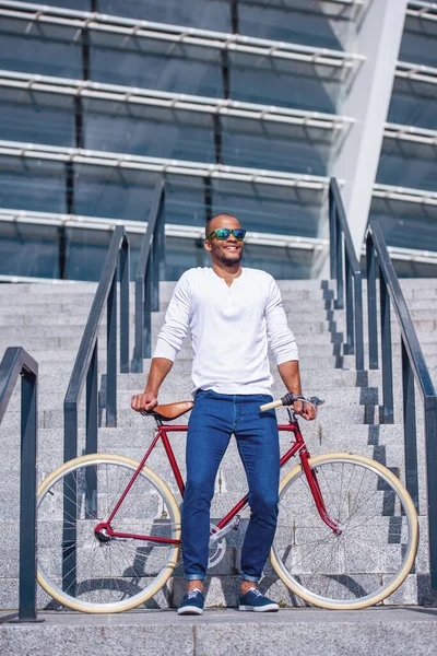 階段の屋外に立っている間 カジュアルな服装と太陽の眼鏡を笑顔と自転車にもたれてハンサムな若いビジネスマンの完全な長さの肖像画 — ストック写真