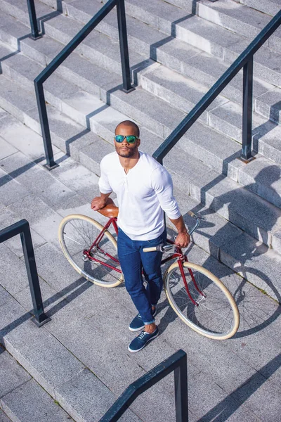 階段の屋外に立っている間に彼の自転車にもたれカジュアルウェアやサングラスでハンサムな若いビジネスマンの高い角度のビュー — ストック写真