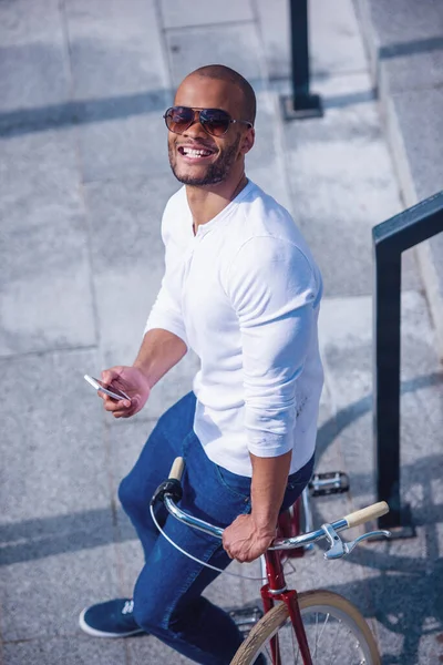 カジュアルな服装のハンサムな若いビジネスマンは スマートフォンを使用しています カメラを見て 彼の自転車にもたれながら笑顔 屋外に立って — ストック写真