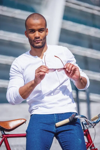 穿着休闲装的年轻貌美的商人拿着太阳镜 看着相机 靠在外面的自行车上 — 图库照片