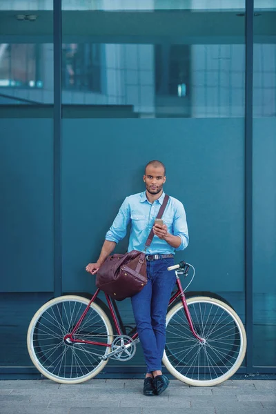 スマートフォンを使い 自転車にもたれながらカメラを眺めながら屋外に立つ カジュアルな服装でハンサムな若いビジネスマンの完全な長さの肖像画 — ストック写真