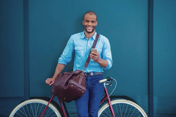 カジュアルな服を着たハンサムな若いビジネスマンは スマートフォンを使用しています カメラを見て 彼の自転車にもたれながら笑顔 屋外に立って — ストック写真