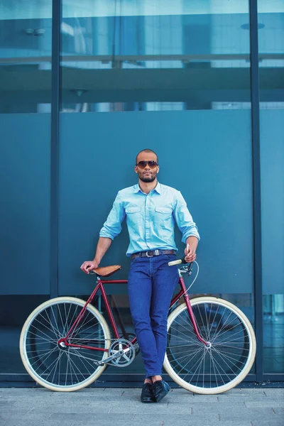 屋外に立って 彼の自転車にもたれながらカメラを見てカジュアルな服やサングラスでハンサムな若いビジネスマンの完全な長さの肖像画 — ストック写真