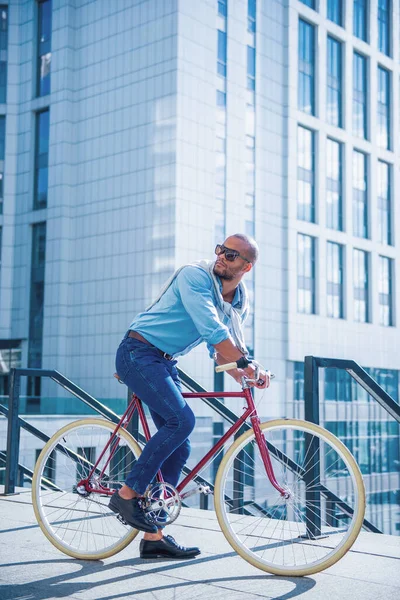 彼の自転車に座って離れて見てカジュアルな服やサングラスでハンサムな若いビジネスマンの完全な長さ — ストック写真