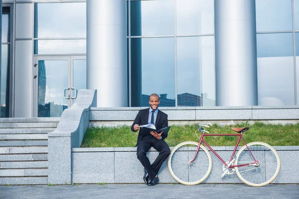 古典的なスーツのハンサムなビジネスマンは 彼の自転車の近くのオフィスビルの外に座っている間 カメラを見て笑って 文書を検討しています — ストック写真