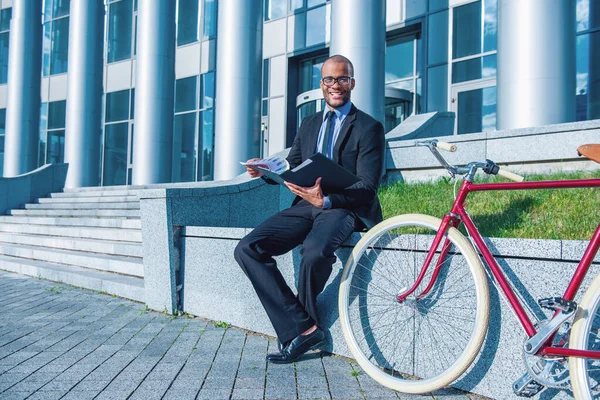 彼の自転車の近くのオフィスビルの外に座って カメラと笑顔を見て 文書を検討しています — ストック写真