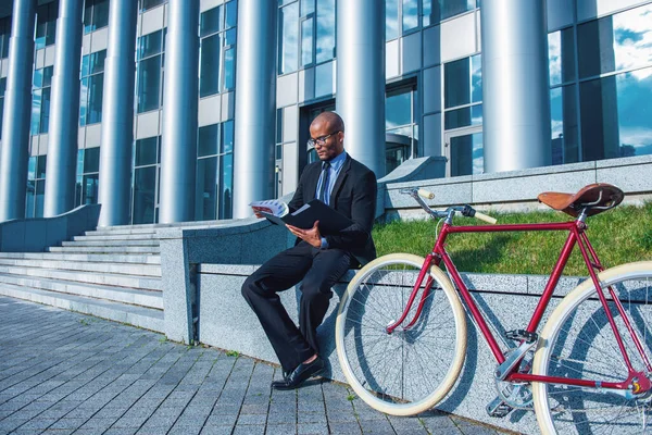古典的なスーツや眼鏡のハンサムなビジネスマンは 彼の自転車の近くのオフィスビルの外に座っている間 文書を調べています — ストック写真