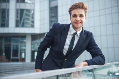 Klasik takım elbiseli çekici genç emin iş adamı kameraya bakıyor ve gülümsüyor, ofis binası dışında balkon korkuluk üzerine eğilerek