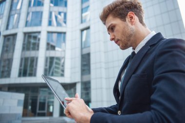 Ofis binası dışında duran bir tablet kullanarak Klasik takım elbiseli çekici genç emin iş adamı yan görünüm