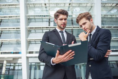 Klasik takım elbiseli iki yakışıklı genç iş adamları bir klasör tutarak ve belgeler, ofis binası dışında ayakta incelenmesi