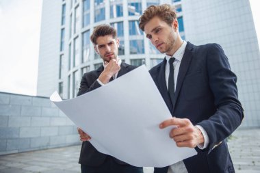 Klasik takım elbiseli iki yakışıklı genç iş adamları kağıt tutan ve ofis binası dışında ayakta belge inceleyerek