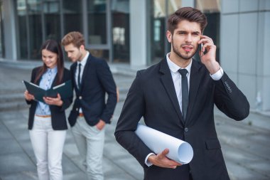 Yakışıklı genç işadamı haddelenmiş kağıt plan ve açık havada duran cep telefonu üzerinde konuşurken tutuyor. İki iş kişi içinde belgili tanımlık geçmiş