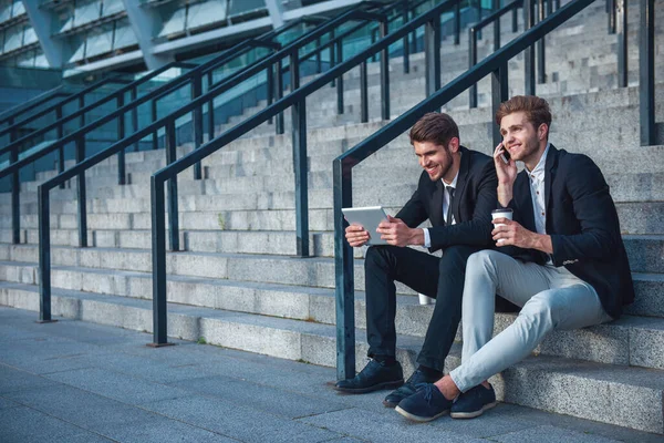 つのハンサムな若いビジネスマンがタブレットを使用して 携帯電話で話しているとコーヒー ブレークの間にオフィスビルの階段に座って笑顔 — ストック写真