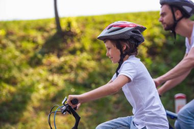 Sevimli küçük çocuk ve babası rahat kıyafetler ve park motosiklet sürme sırasında gülümseyen kask yan görünüm. Baba içinde belgili tanımlık geçmiş
