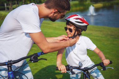 Yakışıklı genç Baba ve sevimli küçük oğlu rahat kıyafetler ve güneş gözlüğü Park'taki bisiklet sürme vardır. Baba, oğlunun Kask sabitleme