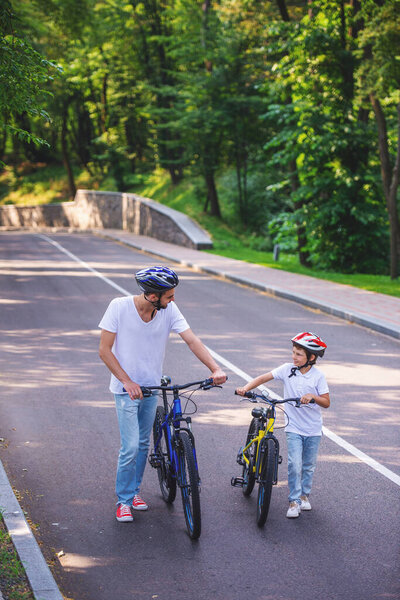 Handsome young dad and his cute little son are looking at each other and smiling while riding bikes in park