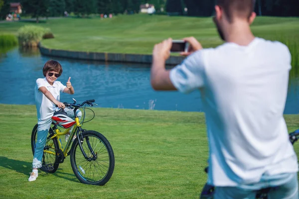 スマート フォンを使用して彼の息子の写真を取っています 小さな男の子は のサインを示す カメラ目線 公園で自転車に座って笑顔 — ストック写真
