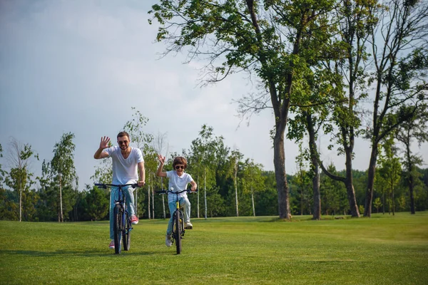 ハンサムな若いお父さんとカジュアルな服とサングラスで彼はかわいい息子が手を振って カメラを見て 公園で自転車に乗りながら笑みを浮かべて — ストック写真