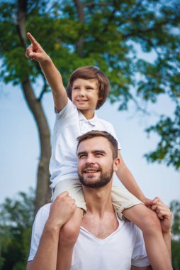 Sevimli küçük bir çocuk ve onun yakışıklı genç Baba parkta dinlenme sırasında ileri ve gülümseyen arıyoruz. Oğlu babasının omuzlarında oturan ve uzağa işaret