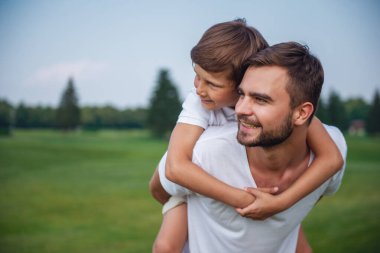 Sevimli küçük bir çocuk ve onun yakışıklı genç Baba parkta dinlenme sırasında ileri ve gülümseyen arıyoruz. Oğlu pickaback oturuyor