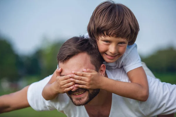 可爱的小男孩和他英俊的年轻爸爸看相机 在公园里休息时微笑的肖像 儿子是坐 Pickaback 和盖在他父亲的眼睛 — 图库照片