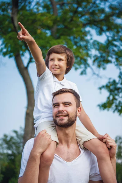 可爱的小男孩和他英俊的年轻爸爸所期待 面带笑容 在公园里休息时 儿子坐在他父亲的肩膀上 指着不远处 — 图库照片