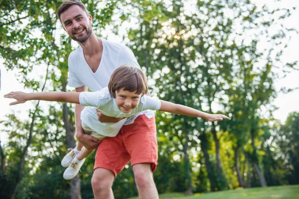 英俊的年轻爸爸和他可爱的小儿子是看相机 微笑着在公园玩 怀里的父亲抱着他的儿子 小男孩就模仿一架飞机 — 图库照片