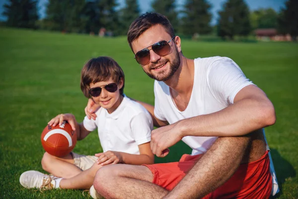 可爱的小男孩和他英俊的年轻爸爸的眼镜孙抱着球看着相机 微笑着坐在公园的草地上 — 图库照片