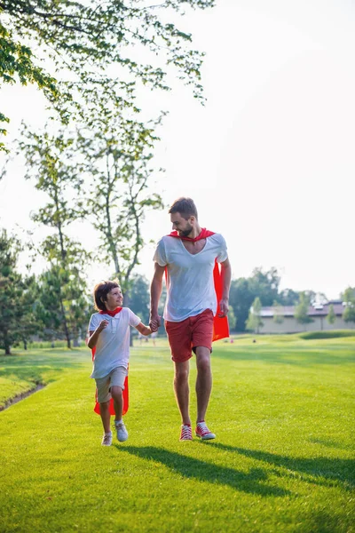 全长肖像的英俊的年轻爸爸和他可爱的小儿子穿得像超级英雄 看着对方 而在公园散步时微笑 — 图库照片