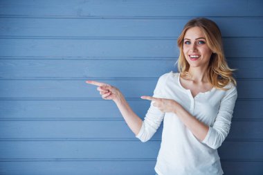 Güzel genç kadın kameraya bakıyor, işaret ediyor ve gülümsüyor, gri duvara karşı duruyor.