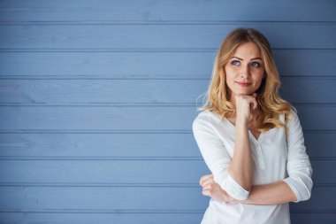 Güzel genç kadın çene üzerinde el tutuyor, uzağa bakıyor ve gülümsüyor, gri duvara karşı ayakta